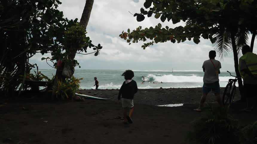 A curious child runs in slow motion towards a tropical black sand beach as spectators watch surfers ride the ocean waves