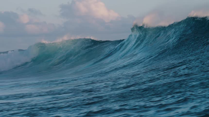 A powerful and magnificent ocean wave swells and barrels in dramatic slow motion under a soft, cloudy sky