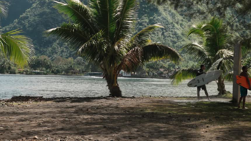 A surfer rinses off at an outdoor shower on a tropical beach while his friend waits nearby with a boogie board