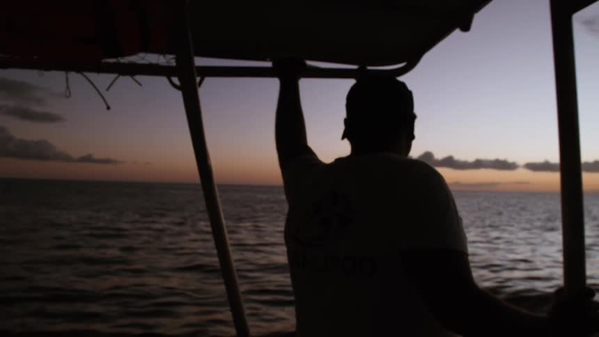 A silhouetted man stands at the helm of a boat, looking out at the calm ocean during a beautiful sunset
