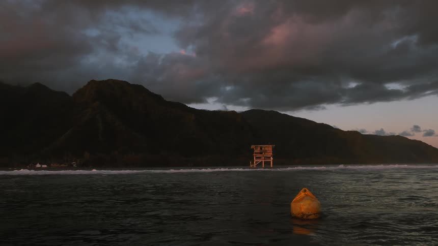 A yellow buoy floats in the ocean at dusk with stormy clouds gathering over a mountainous island and a surf competition tower on the shore