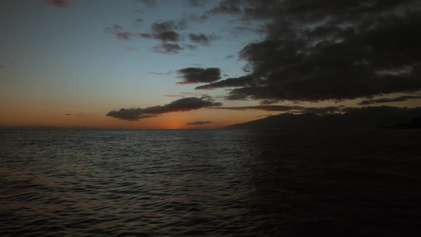 The sun dips below the horizon over the calm sea, under a sky filled with dark, moody clouds