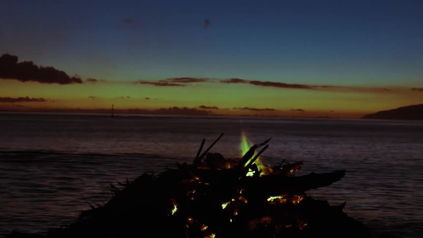 A tranquil bonfire burns on the beach next to the calm ocean as the sun sets over the horizon