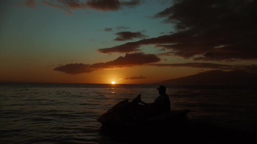 A silhouetted person sits on a jet ski in the calm ocean, watching a picturesque sunset over a distant island