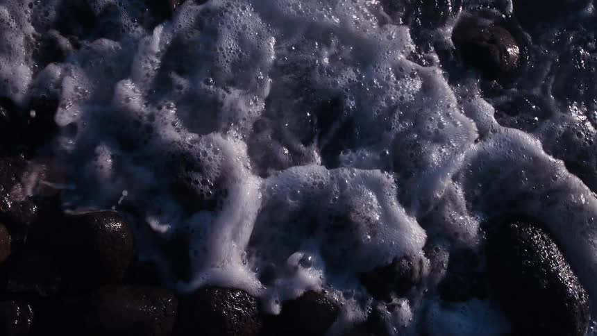A slow-motion overhead view of foamy ocean waves gently washing over dark, wet rocks on the shoreline