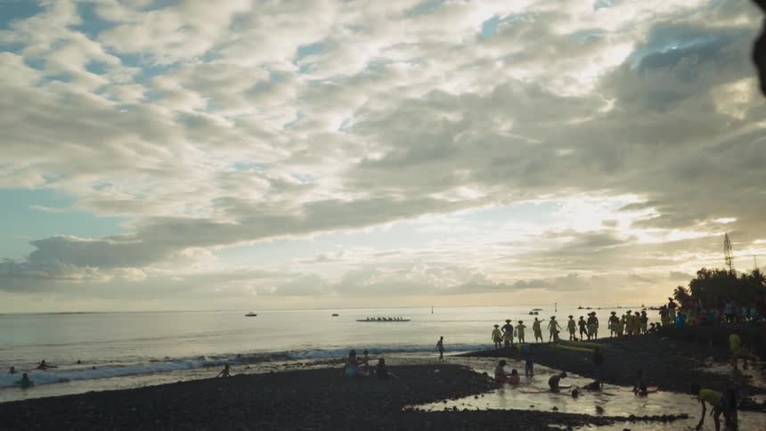 Silhouetted crowds cheer on an outrigger canoe team from the shore of a tropical beach during a golden sunset