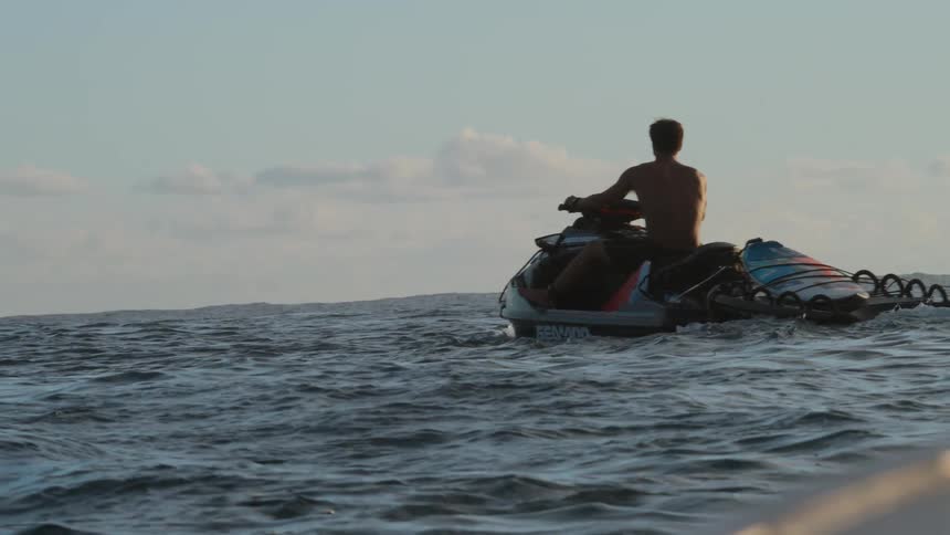 A lone surfer on a jet ski waits patiently in the open ocean with his board, silhouetted against the golden hour sky