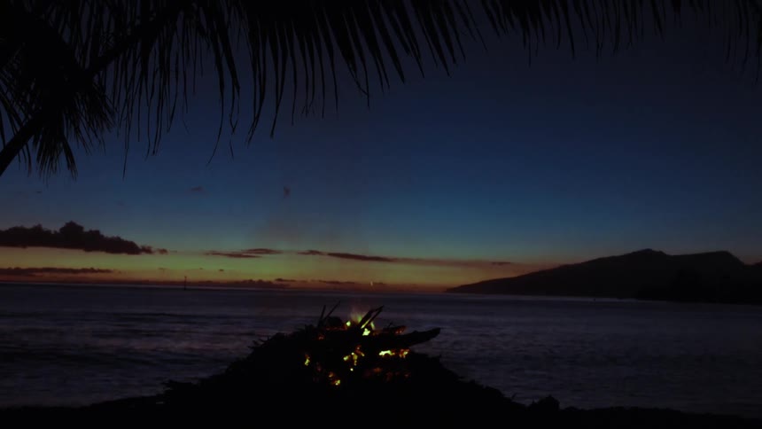 A serene bonfire crackles on a tropical beach as the last light of sunset fades over the ocean and a distant island