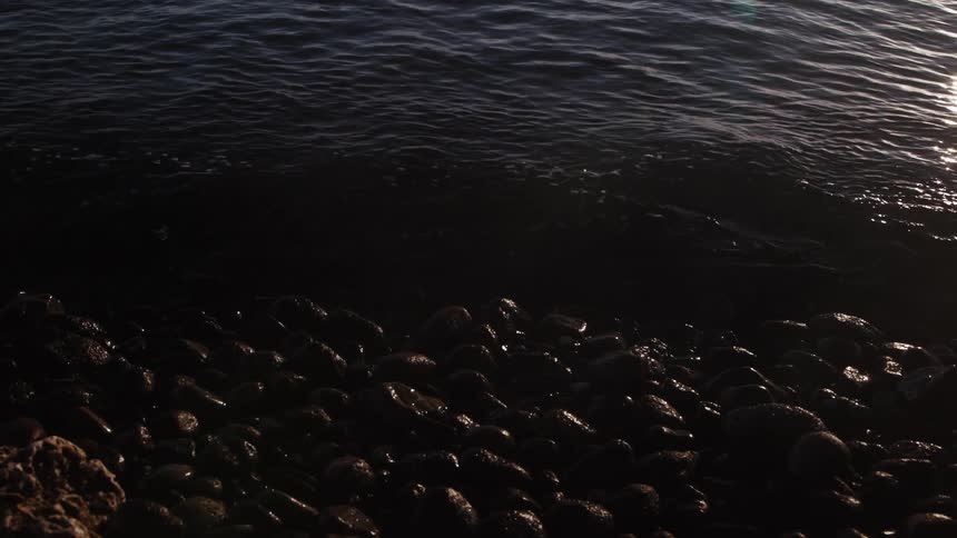 Calm ocean waves gently wash over a rocky beach in slow motion with the setting sun reflecting on the water