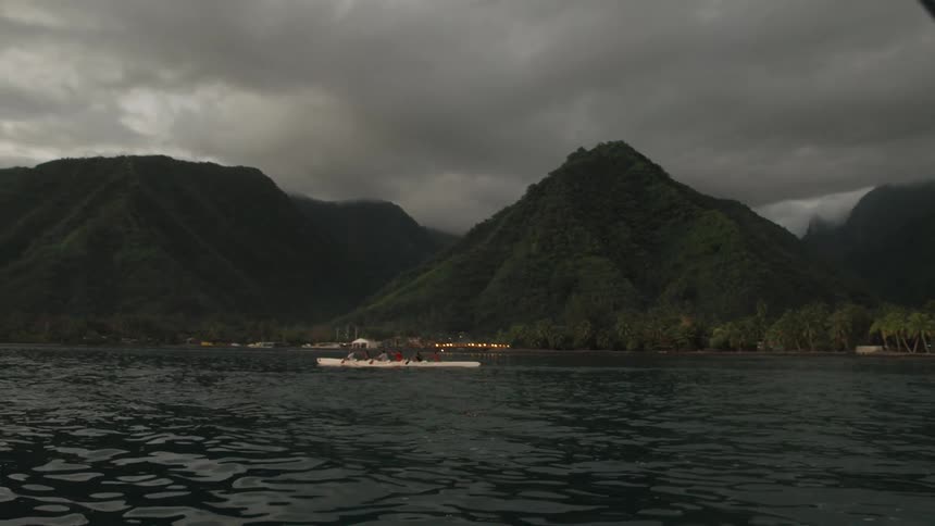 A team of rowers in an outrigger canoe paddle through calm waters along a tropical island with lush mountains under a dramatic, cloudy sky
