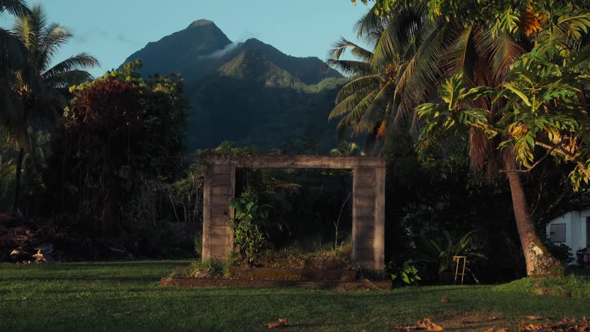 A weathered stone gateway sits in a tranquil tropical yard, framing a view of a towering green mountain at dawn