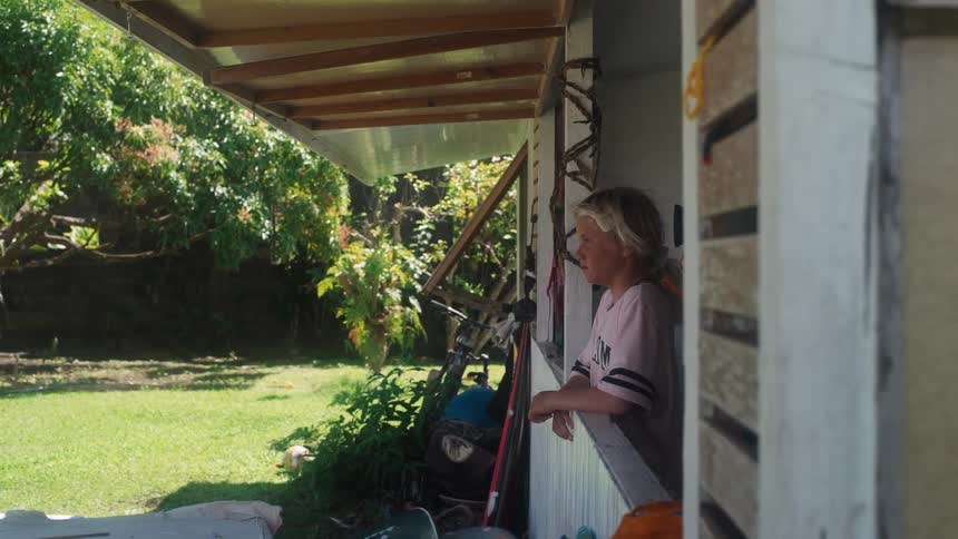 A thoughtful young girl with blonde hair leans on an open window on her porch, looking out at the lush green yard on a sunny day