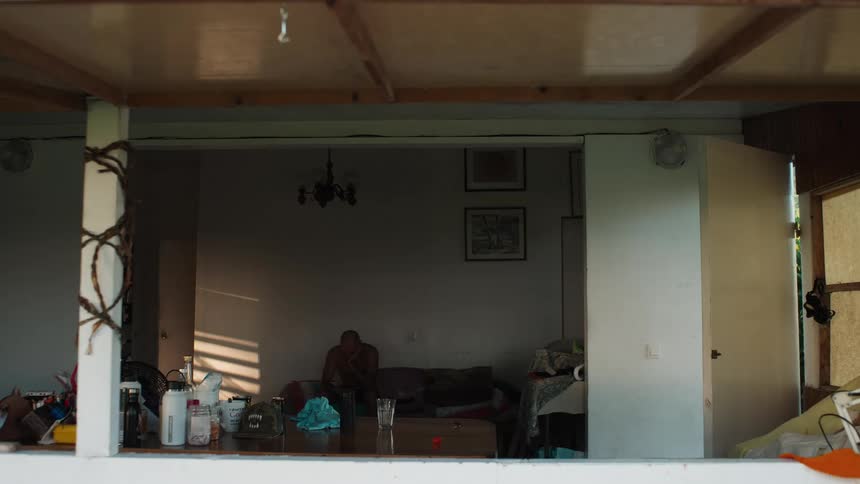 A shirtless man sits alone in quiet contemplation on a couch in a rustic room as sunlight casts long shadows on the wall