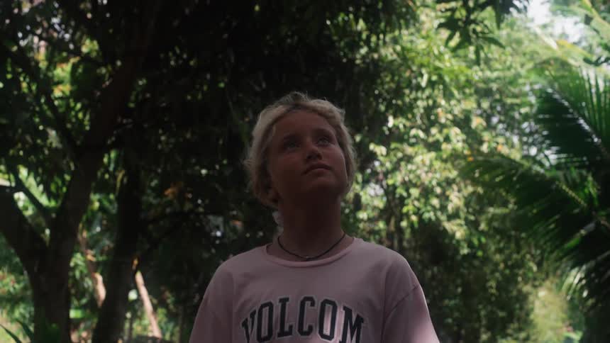 A young blonde-haired person looks up in awe amidst the dense green foliage of a tropical jungle