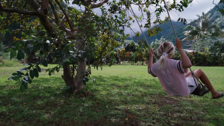 Slow motion view of a blonde girl swinging on a rope swing under a tree, with lush green mountains in the distance
