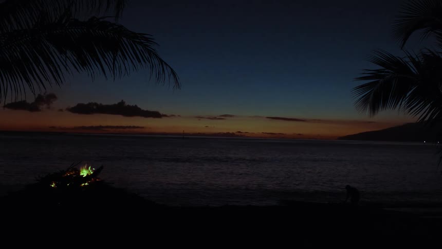A tranquil tropical evening unfolds with a glowing bonfire on the beach and a silhouetted person by the ocean's edge at dusk