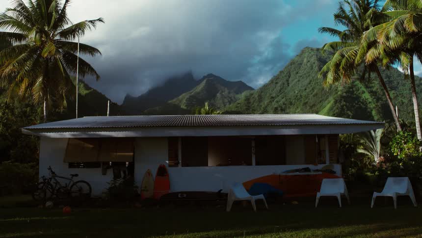 A tranquil surf shack sits nestled amongst palm trees with a dramatic mountain landscape in the background