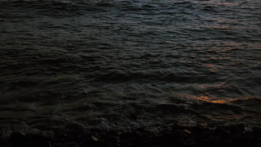 A slow upward tilt from a dark, rocky shore reveals a stunning golden sunset over the ocean