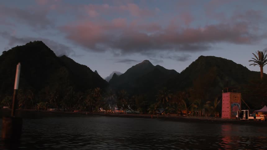 A view from the water of a tropical beach village at dusk, nestled at the base of lush mountains