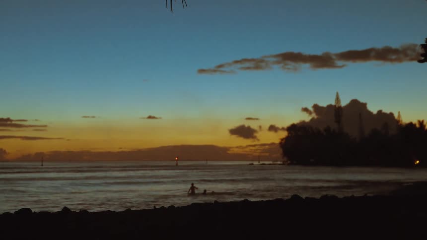 Palm fronds frame a beautiful tropical sunset as silhouetted people enjoy a peaceful swim in the calm ocean