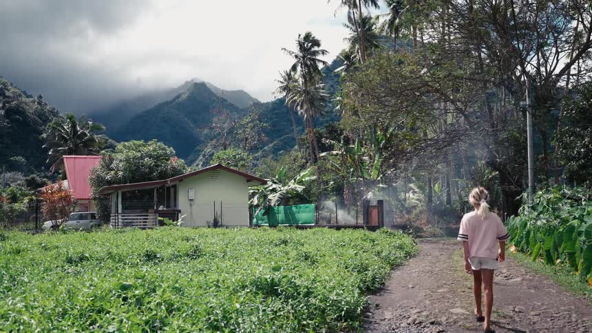 A blonde woman walks down a dirt road in a rural tropical village with lush green fields and misty mountains in the background