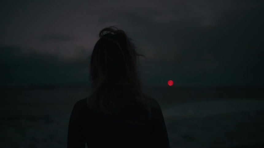A lone female surfer stands in the dark, contemplating the ocean waves after sunset