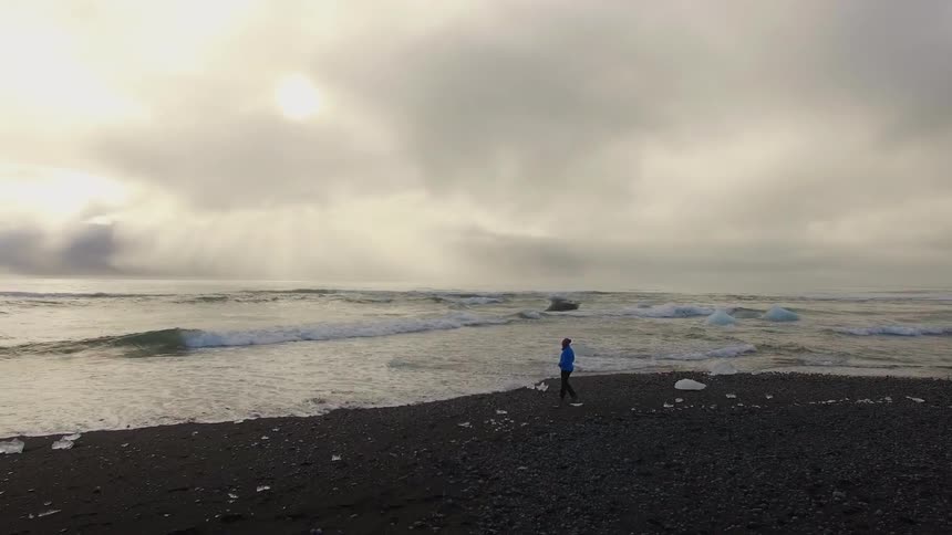 A person in a coat walks the black sand beach as waves come to shore