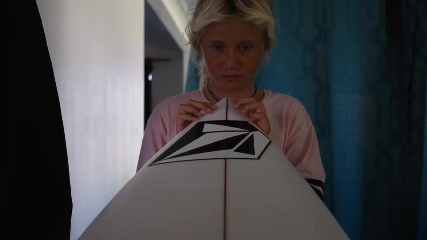 A young blonde surfer stares intensely down the tip of their surfboard in a dramatic slow-motion shot