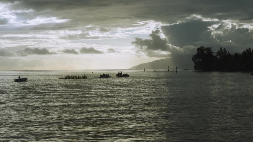 A team of silhouetted paddlers in an outrigger canoe rows across a calm ocean under a dramatic, cloudy sky at sunrise