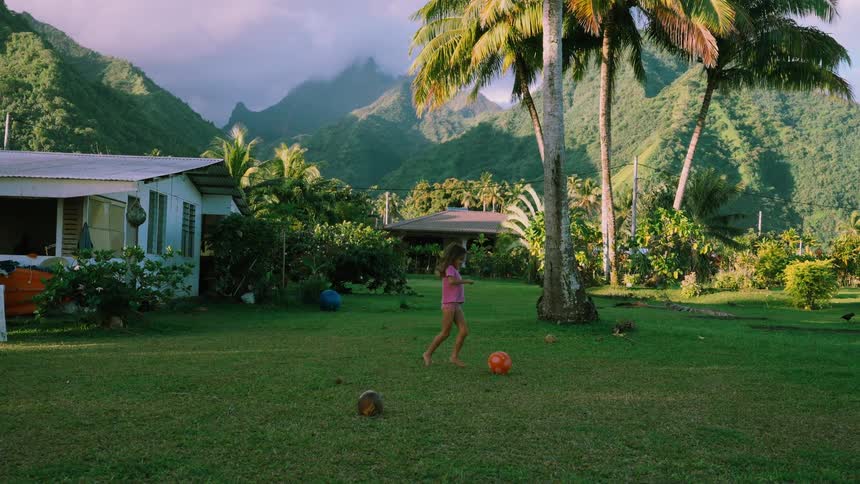 A young barefoot girl with curly hair plays with a ball in slow motion in a lush tropical yard surrounded by palm trees and majestic green mountains