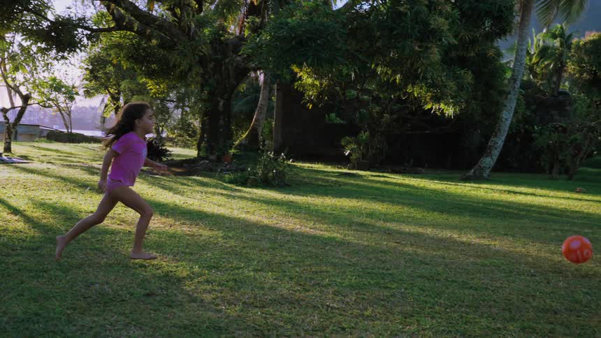 A young girl with long curly hair runs barefoot across a sunlit green lawn, chasing a red ball in slow motion
