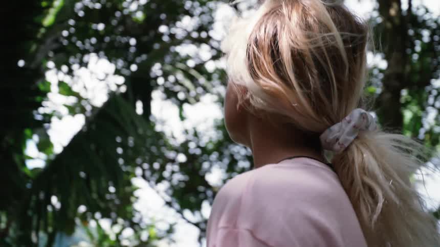 A young blonde woman gazes up with wonder at the sunlit canopy of a lush tropical jungle in slow motion
