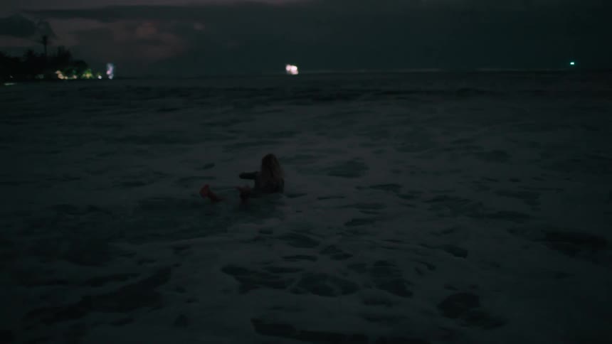 A female surfer walks into the dark ocean at dusk with her surfboard and begins to paddle out into the waves