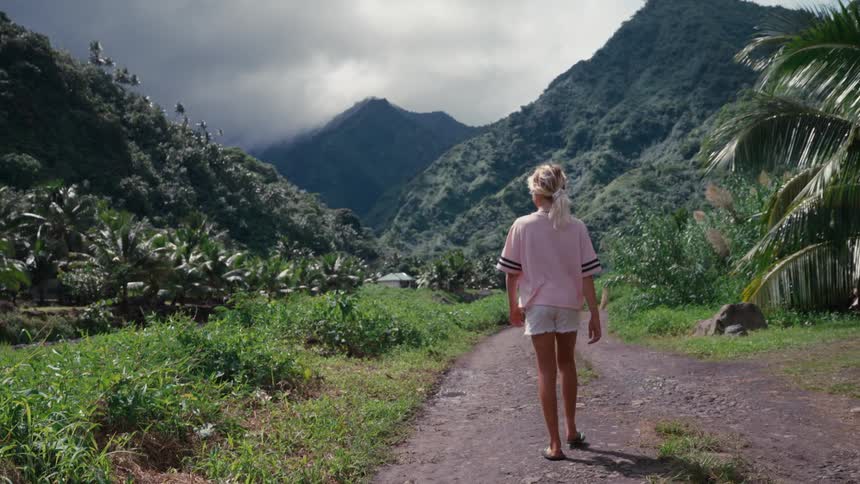 A young blonde woman leisurely walks down a remote dirt path, heading into a stunning tropical mountain valley