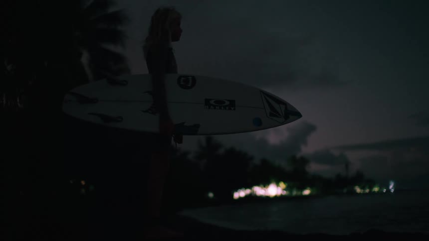 A silhouetted surfer stands with their board on a tropical beach at night, gazing at the distant lights across the water