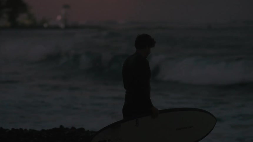 A silhouetted surfer in a wetsuit holds his surfboard while watching the ocean waves at dusk in slow motion