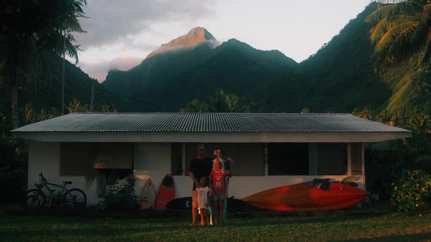 A beautiful family portrait in front of a modest island home at dusk with majestic green mountains in the background