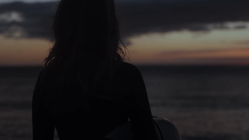 A silhouetted female surfer in a wetsuit holds her board and watches the ocean waves at sunset