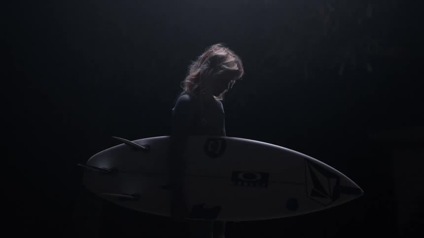 A surfer stands pensively in the dark holding a surfboard, illuminated by a single spotlight