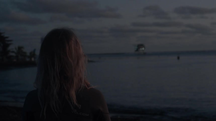 A blonde woman in a wetsuit stands on a dark beach at twilight, looking out at the calm sea