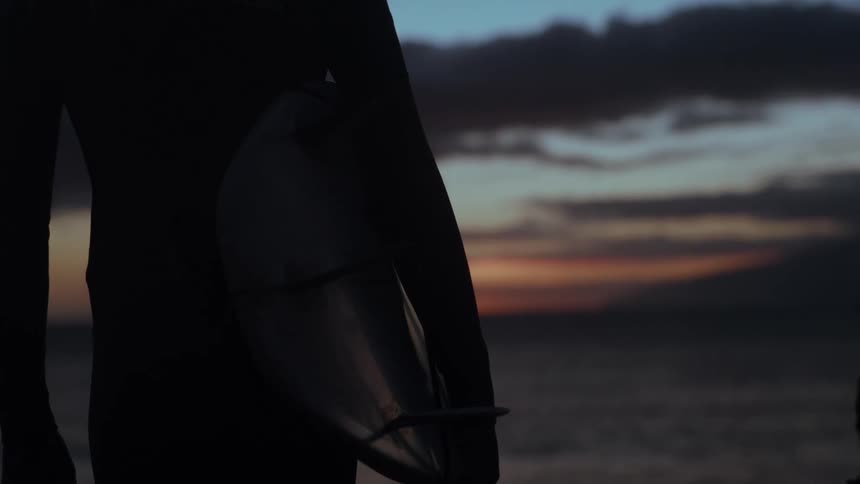 Close up of a silhouetted surfer holding a surfboard and looking out at the ocean at dusk