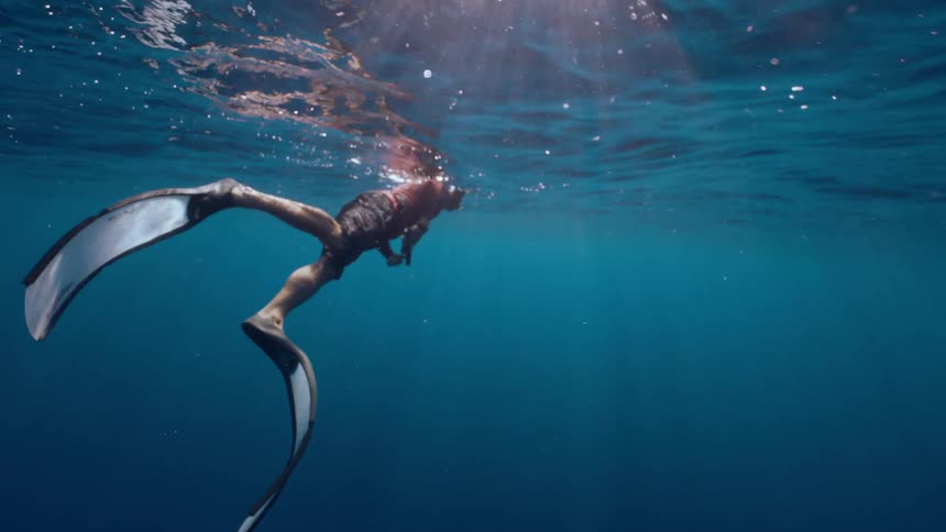 An underwater shot shows a man in freediving fins floating near the surface of the deep blue ocean with sunbeams shining through