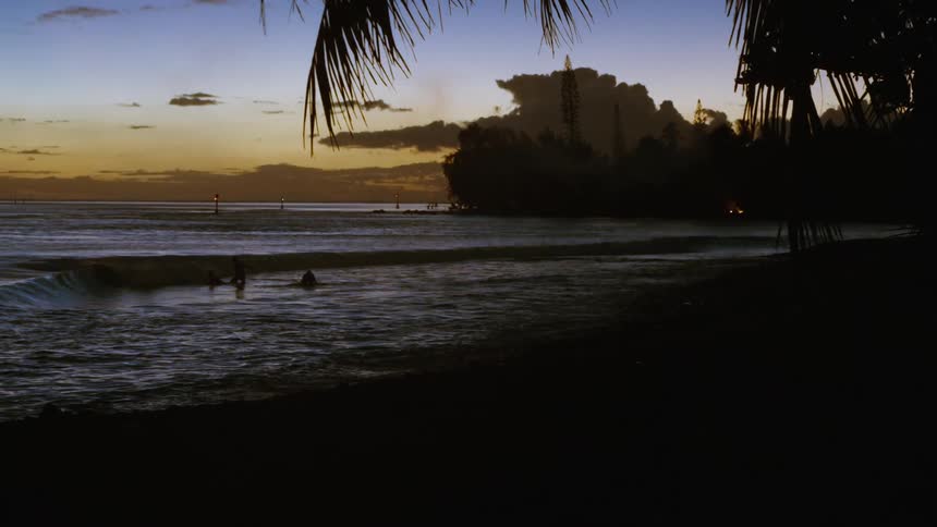 Silhouetted surfers enjoy the last light of the day as gentle waves roll onto a tropical beach at dusk