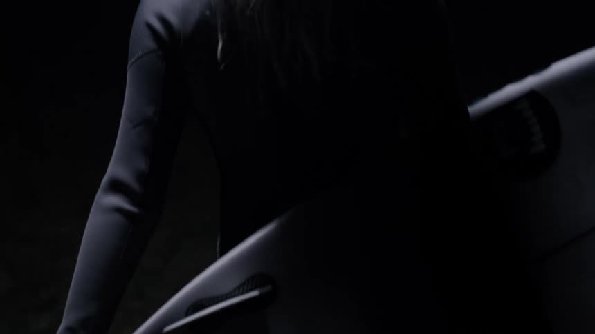 A slow-motion close-up reveals a female surfer in a wetsuit holding her board in the dark with dramatic lighting