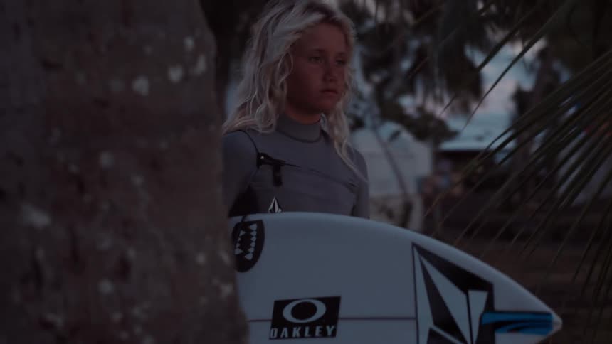 A focused young surfer with blonde hair holds their surfboard, standing amongst palm trees at twilight