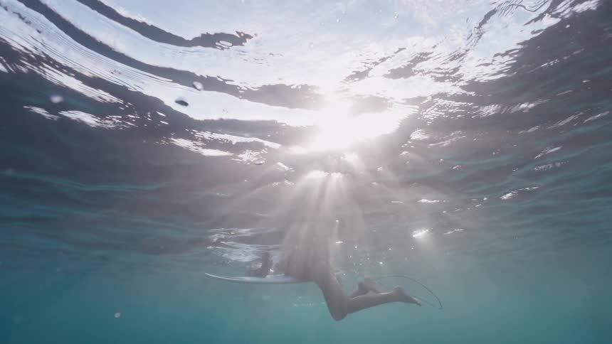 A serene underwater shot captures a surfer floating on their board, illuminated by brilliant sun rays from above
