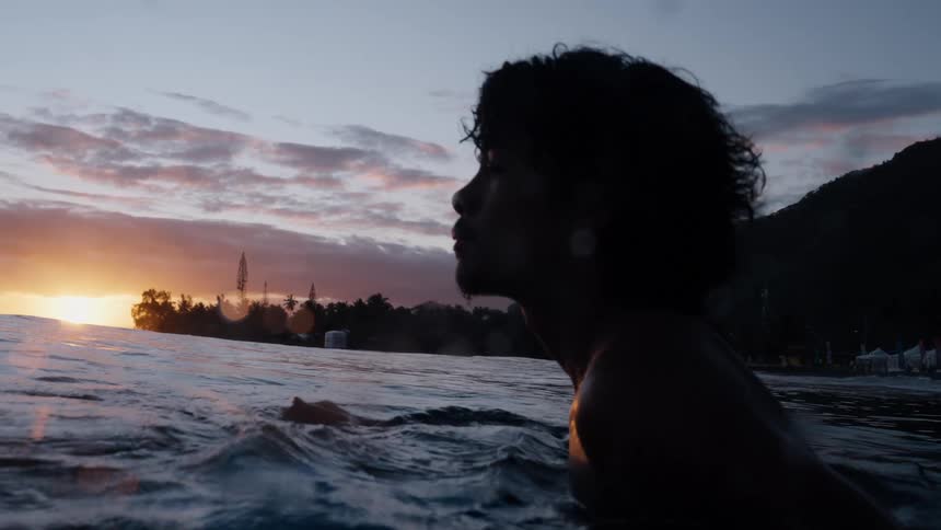 A silhouetted surfer with curly hair waits in the ocean for a wave as the sun sets in slow motion