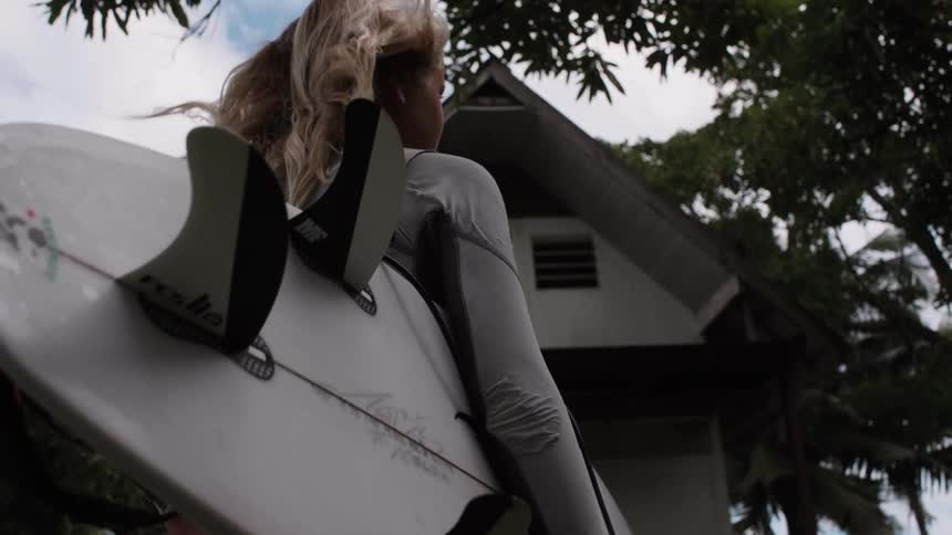 A female surfer in a wetsuit holds her surfboard as her hair blows in the wind in slow motion