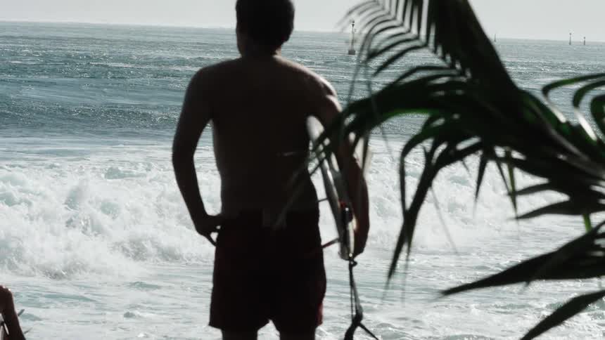 A silhouetted surfer holding his board stands at the water's edge, watching the waves roll in