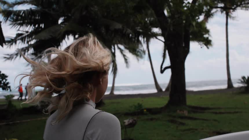 Slow motion shot from behind of a female surfer with blonde hair blowing in the wind as she carries her surfboard towards the tropical ocean